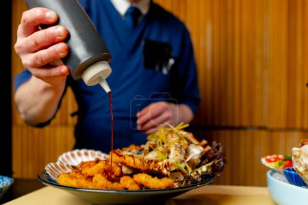 Téléchargez les photos : Gros plan dans un restaurant japonais un chef en uniforme bleu verse de la sauce sur une assiette de fruits de mer - en image libre de droit