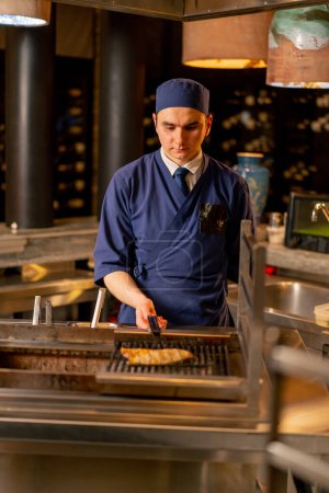 Foto de En un restaurante japonés un chef en uniforme azul fríe pescado en la parrilla de la cocina - Imagen libre de derechos