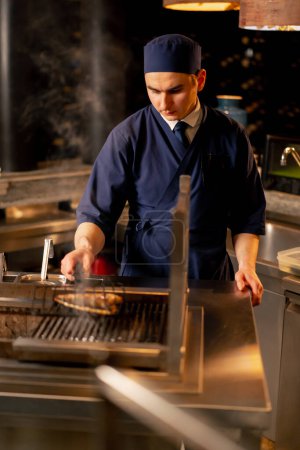 Téléchargez les photos : Dans un restaurant japonais un chef en uniforme bleu frites poisson sur le gril dans la cuisine - en image libre de droit