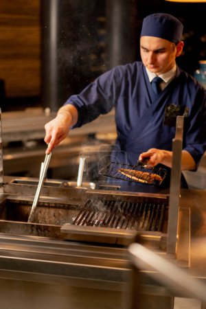 Téléchargez les photos : Dans un restaurant japonais un chef en uniforme bleu frites poisson sur le gril dans la cuisine - en image libre de droit