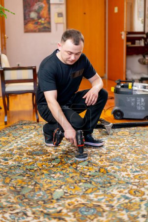 Téléchargez les photos : Dans l'appartement, un maître nettoyeur nettoie le tapis avec du détergent à l'aide d'un tournevis et d'une brosse - en image libre de droit