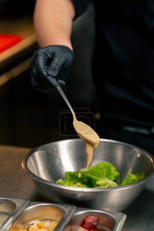 Photo for Close-up in a professional kitchen the chef pours sauce over lettuce leaves in an iron bowl - Royalty Free Image