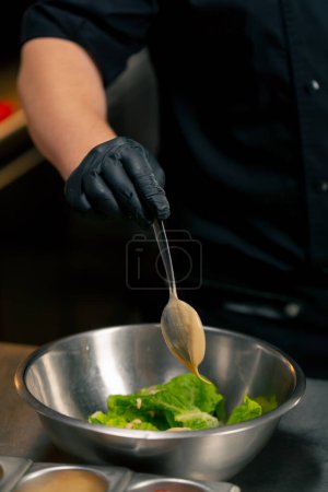 Foto de Primer plano en una cocina profesional el chef vierte salsa sobre las hojas de lechuga en un tazón de hierro - Imagen libre de derechos