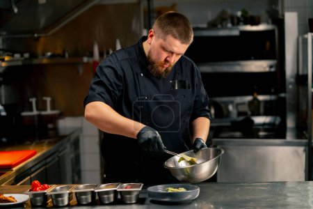 Téléchargez les photos : Dans une cuisine professionnelle, le chef met des morceaux de laitue en sauce dans une assiette avec une pince à épiler - en image libre de droit
