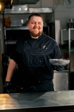 Téléchargez les photos : Dans la cuisine professionnelle, le chef se tient dans ses mains avec une salade préparée et sourit à la caméra - en image libre de droit