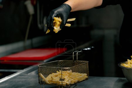 Téléchargez les photos : Close-up dans un chef de cuisine professionnel en gants noirs met des frites dans une friteuse - en image libre de droit