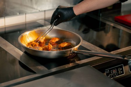 Foto de Primer plano en una cocina profesional las alas de pollo se fríen en sartén con salsa que el chef revuelve con pinzas - Imagen libre de derechos