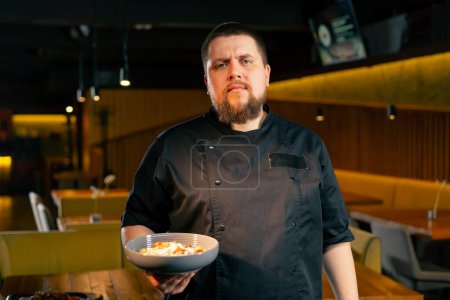 Téléchargez les photos : Dans un chef de restaurant dans une veste noire se tient avec une salade préparée en regardant la caméra - en image libre de droit
