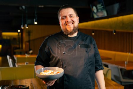 Téléchargez les photos : Dans un restaurant un chef dans une veste noire se tient avec un sourire de salade préparé regarde la caméra - en image libre de droit
