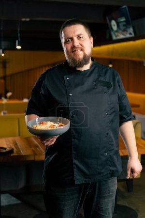 Téléchargez les photos : Dans un restaurant un chef dans une veste noire se tient avec un sourire de salade préparé regarde la caméra - en image libre de droit