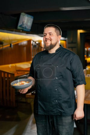 Téléchargez les photos : Dans un restaurant chef dans une veste noire se tient souriant avec une salade préparée - en image libre de droit