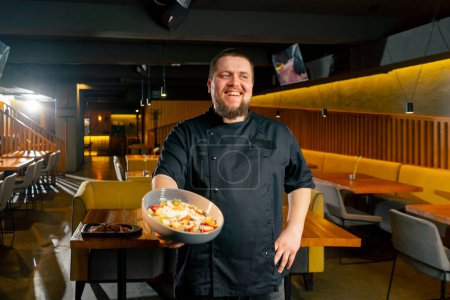 Téléchargez les photos : Dans un restaurant chef dans une veste noire se tient souriant avec une salade préparée - en image libre de droit