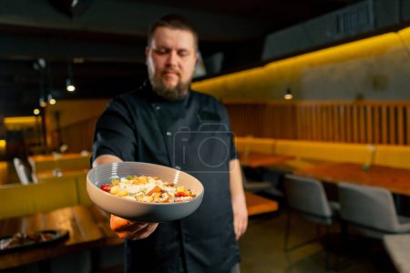 Téléchargez les photos : Dans un chef de restaurant dans une veste noire se tient avec une salade préparée en regardant la caméra - en image libre de droit
