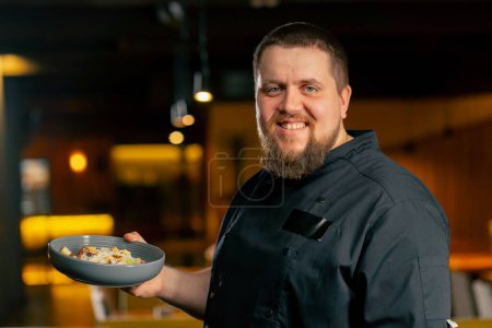 Téléchargez les photos : Gros plan dans un chef de restaurant dans une veste noire se tient sourire avec une salade préparée - en image libre de droit