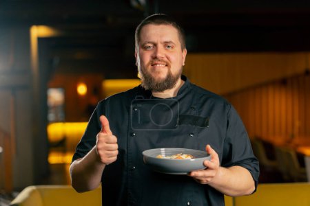 Téléchargez les photos : Gros plan dans un chef de restaurant dans une veste noire se tient souriant avec une salade préparée super doigt - en image libre de droit