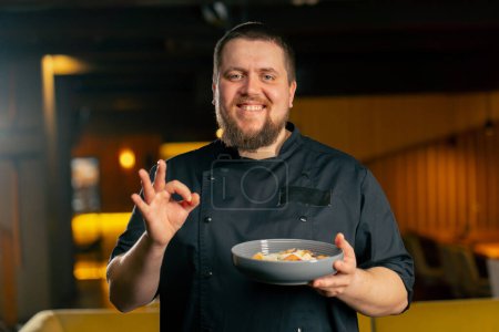 Téléchargez les photos : Gros plan dans un chef de restaurant dans une veste noire se tient souriant avec une salade préparée super doigt - en image libre de droit