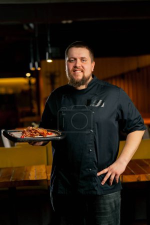 Téléchargez les photos : Dans un chef de restaurant dans une veste noire se tient souriant avec une préparation ailes de poulet - en image libre de droit