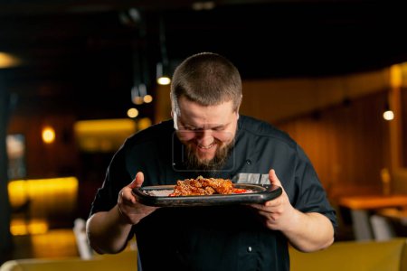 Téléchargez les photos : Fermer un chef en noir dans un restaurant renifle les ailes de poulet qu'il a lui-même préparé - en image libre de droit