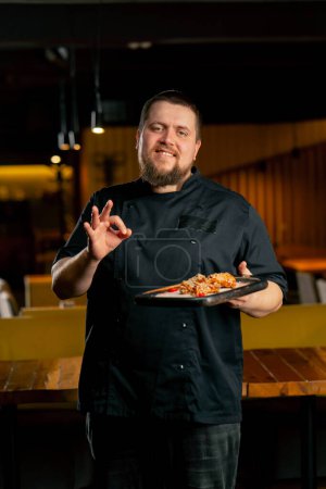 Téléchargez les photos : Dans un chef de restaurant dans une veste noire se tient souriant avec un poulet préparé ailes montrer super doigt - en image libre de droit