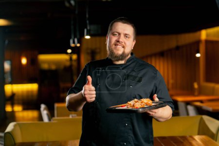 Téléchargez les photos : Dans un chef de restaurant dans une veste noire se tient souriant avec un poulet préparé ailes montrer super doigt - en image libre de droit