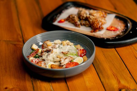 Téléchargez les photos : Close-up sur une table en bois il y a un plat préparé de salade césar dans une assiette grise et des ailes de poulet - en image libre de droit