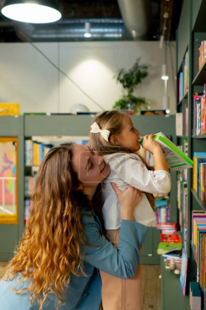 Téléchargez les photos : Dans une librairie dans la zone enfants une mère aide sa fille à atteindre le bon livre - en image libre de droit