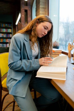 Photo for In a bookstore near the window a beautiful student is sitting reading books preparing for exams - Royalty Free Image
