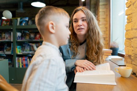 Téléchargez les photos : Fermer dans une librairie près de la fenêtre une jeune mère s'assoit pour apprendre à son fils à lire le développement de l'enfant - en image libre de droit