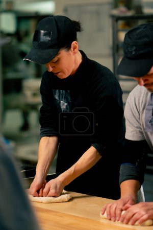 Foto de En una cocina profesional un panadero femenino en una túnica negra forma la forma del pan en una bandeja para hornear - Imagen libre de derechos