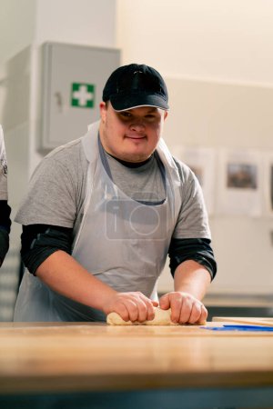 Foto de En una cocina profesional un trabajador inclusivo en uniforme blanco despliega la masa y sonríe - Imagen libre de derechos