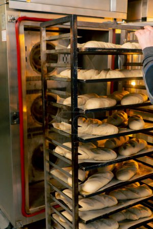 Foto de En una cocina profesional un panadero inclusivo abre el horno y lleva un estante con pan - Imagen libre de derechos