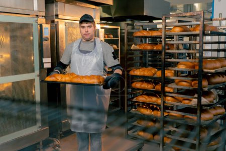 Foto de En la cocina profesional un panadero inclusivo se para con pan listo en una bandeja sonriendo - Imagen libre de derechos