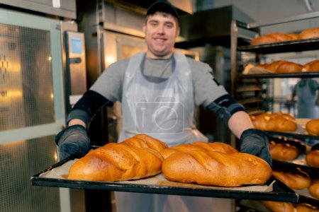 Foto de Primer plano En la cocina profesional un panadero inclusivo se para con pan listo en una bandeja sonriendo - Imagen libre de derechos