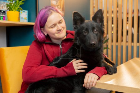 Photo for A young girl with pink hair cuddling with a black dog Belgian Shepherd in a cafe love to animal - Royalty Free Image