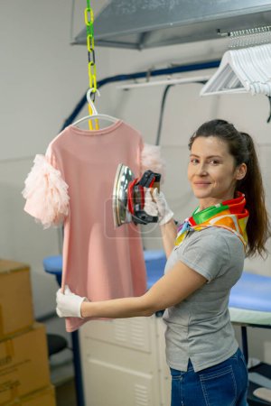 Photo for Professional dry cleaning young girl irons a pink sweater on a hanger with steam look on camera - Royalty Free Image