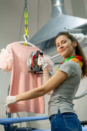 Photo for Professional dry cleaning young girl irons a pink sweater on a hanger with steam look on camera - Royalty Free Image
