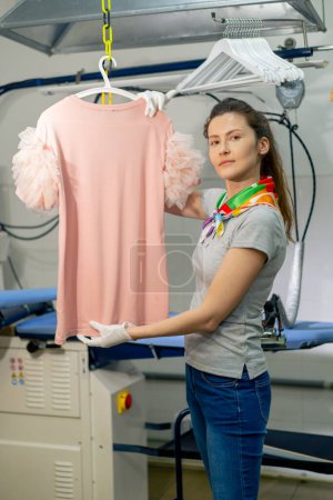 Téléchargez les photos : Professional dry cleaning young girl irons a pink sweater on a hanger with steam look on camera - en image libre de droit