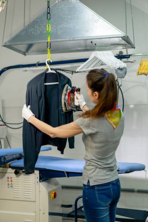 Téléchargez les photos : Professional dry cleaning young girl irons a black sweater on a hanger with steam - en image libre de droit