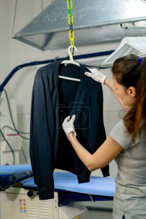 Photo for Professional dry cleaning young girl irons a black sweater on a hanger with steam - Royalty Free Image