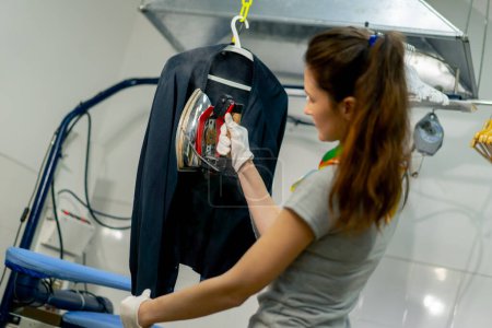 Téléchargez les photos : Professional dry cleaning young girl irons a black sweater on a hanger with steam - en image libre de droit