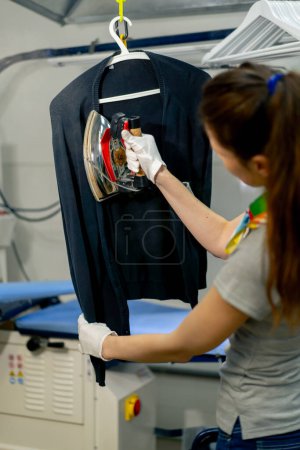 Photo for Professional dry cleaning young girl irons a black sweater on a hanger with steam - Royalty Free Image