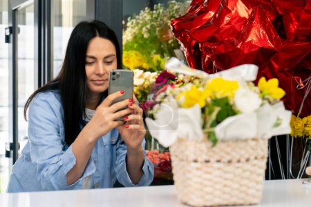 Photo for In a flower shop a florist takes a photo of a collected yellow bouquet in a basket on his phone - Royalty Free Image