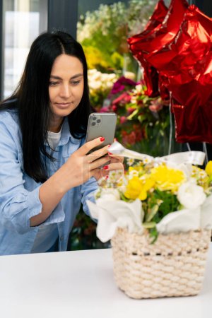 Photo for In a flower shop a florist takes a photo of a collected yellow bouquet in a basket on his phone - Royalty Free Image