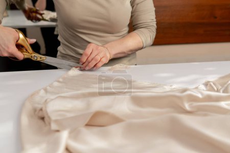 Photo for Close up A professional seamstress cuts cream silk fabric with scissors for a dress custom product - Royalty Free Image