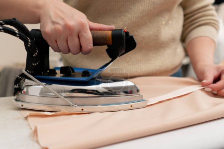 Photo for Close-up in a sewing workshop a woman irons beige fabric with a professional iron - Royalty Free Image