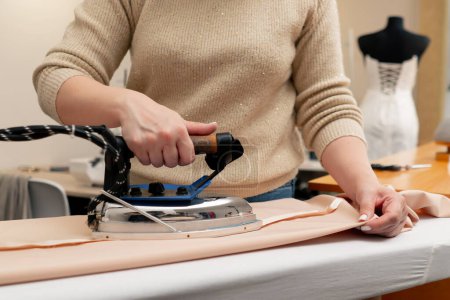 Photo for Close-up in a sewing workshop a woman irons beige fabric with a professional iron - Royalty Free Image