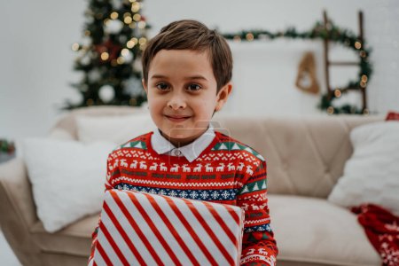 Téléchargez les photos : Mignon petit garçon tenant boîte cadeau et posant à la caméra dans le salon avant Noël. Joyeux Noël et cadeau concept - en image libre de droit