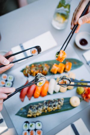 Téléchargez les photos : Friends hands holding sushi and rolls with wooden chopsticks in the restaurant. Japanese and Chinese cuisine concep - en image libre de droit