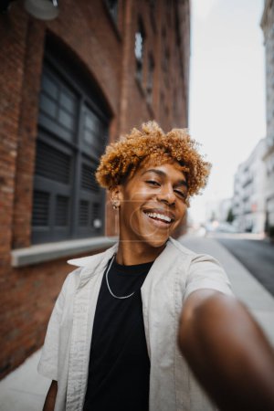 teenager multiracial man smiling confident while making selfie by the camera at the street. Guy with makeup using smartphone camera outdoors in city street. People and photography concept 