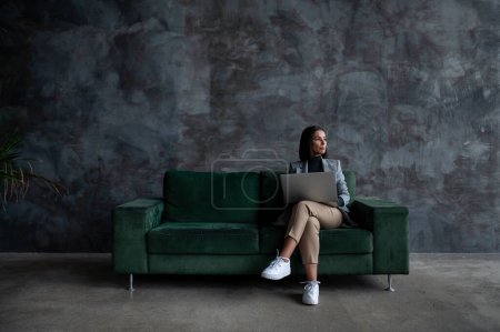 Téléchargez les photos : Jeune créatrice assise sur un canapé vert dans le salon loft et dessinant sur tablette. Femme créative préparant seul un projet d'architecture - en image libre de droit